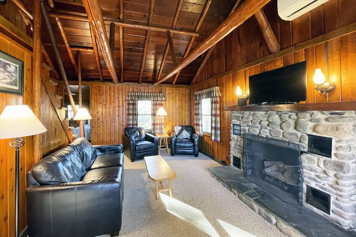 A log cabin interior with a stone fireplace, leather sofas, and a flat-screen TV mounted above the fireplace.