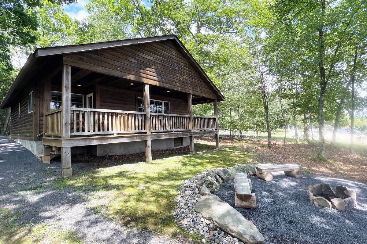 A wooden cabin with a porch, surrounded by trees and a gravel area with logs and a fire pit.
