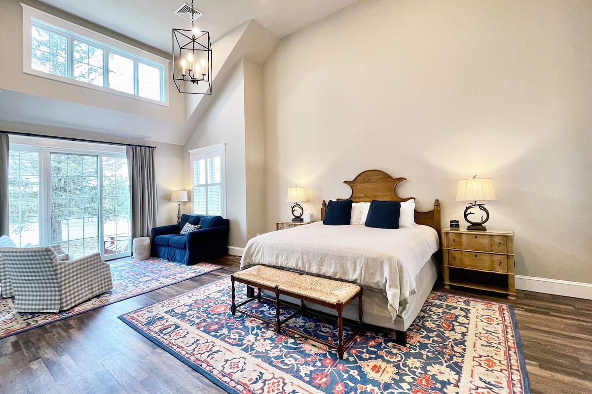 A spacious bedroom with a large bed, a bench, a blue armchair, and a nightstand with a lamp.