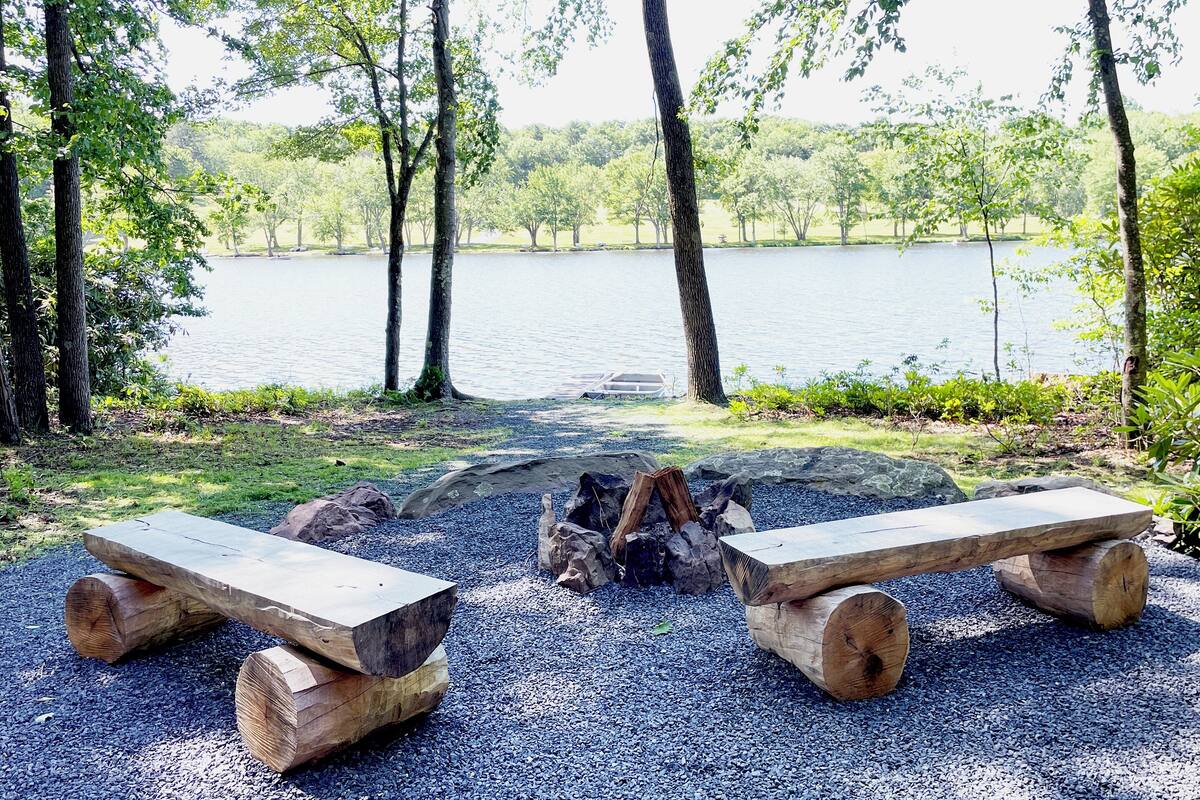 A serene lakeside setting with a fire pit and two wooden benches.