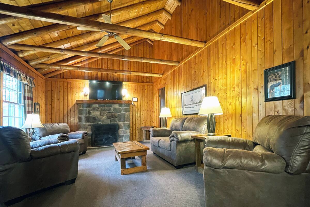 A cozy log cabin interior with a stone fireplace, leather sofas, and a wooden ceiling.