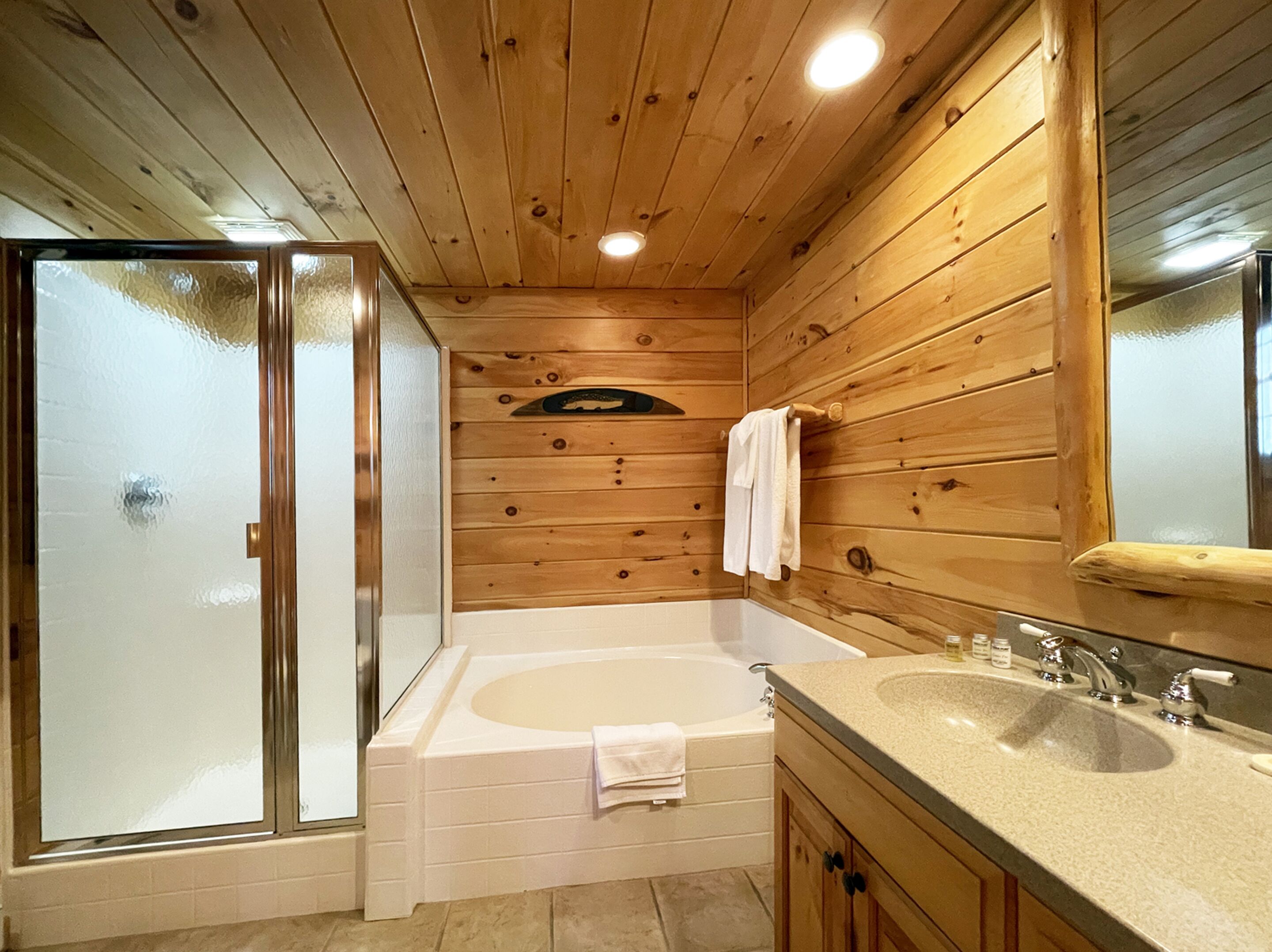 A wooden cabin bathroom with a glass shower enclosure, a bathtub, and a sink with a mirror.