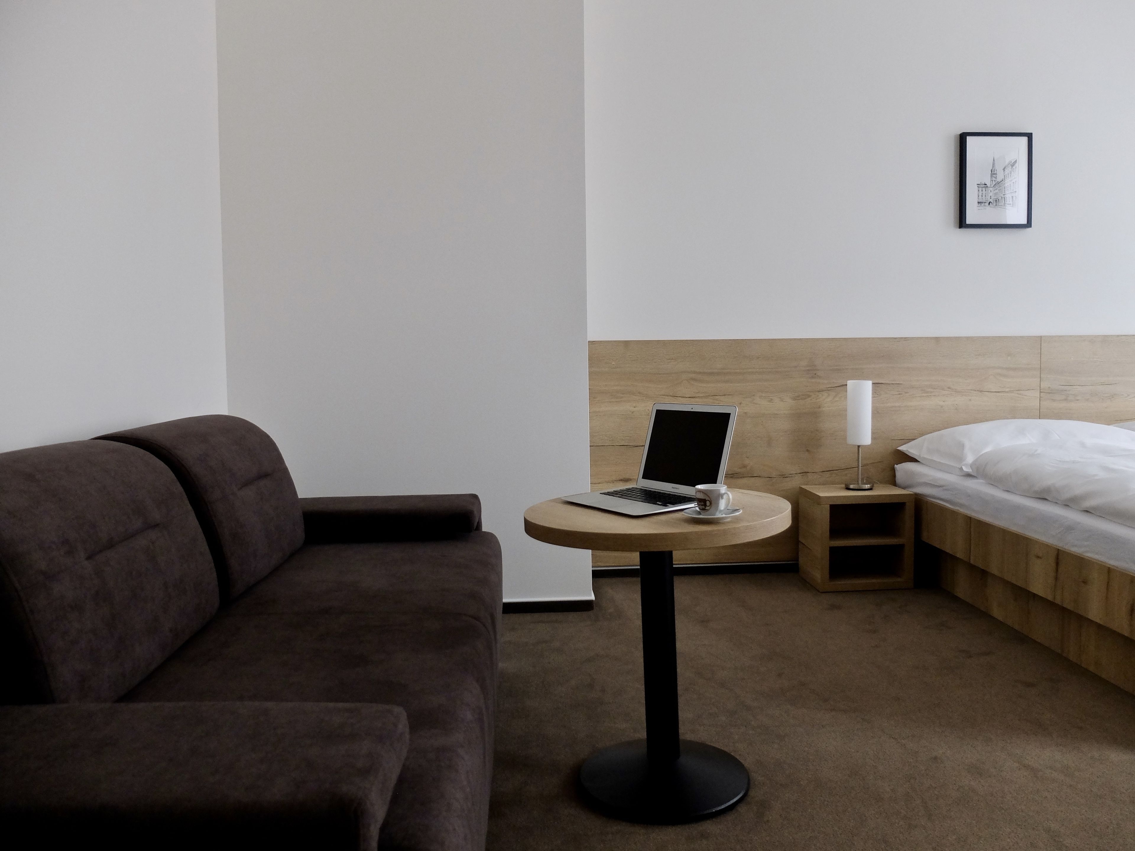 Habitación de hotel moderna con un sofá marrón, una pequeña mesa redonda con una laptop y una taza, una cama con cabecera de madera, y un cuadro enmarcado en la pared.