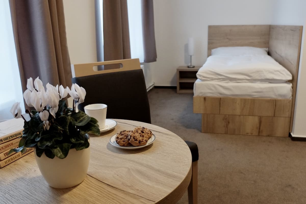 Una habitación de hotel con cama, una mesita de noche con lámpara, una silla, una mesa con planta y galletas, y una ventana con cortinas.