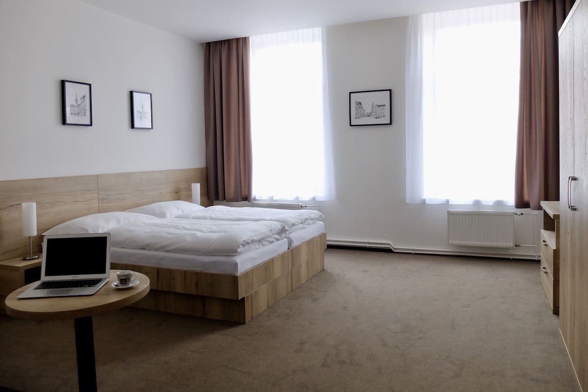 Habitación de hotel con cama de cabecera de madera, mesita de noche con una computadora portátil y una taza, dos cuadros en la pared y ventanas amplias con cortinas.