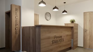 Un comptoir d’accueil avec un revêtement en bois et l’inscription « Pension Stará Posta ». Trois lampes suspendues sont fixées au-dessus du comptoir, et une horloge est visible sur le mur, à gauche.