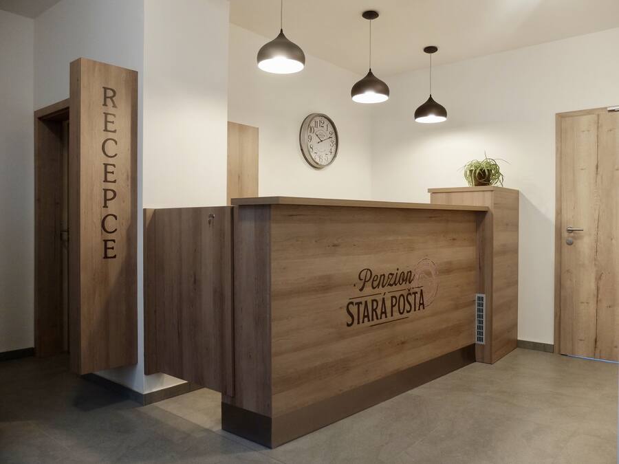 A reception desk with a wooden finish and the text "Pension Stará Posta" on it. There are three pendant lights above the desk, and a clock is visible on the wall to the left.