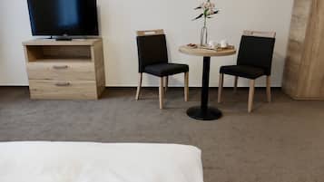 A hotel room with a bed, a TV on a wooden stand, two black chairs around a small round table with a vase of flowers, and a wooden cabinet with drawers.