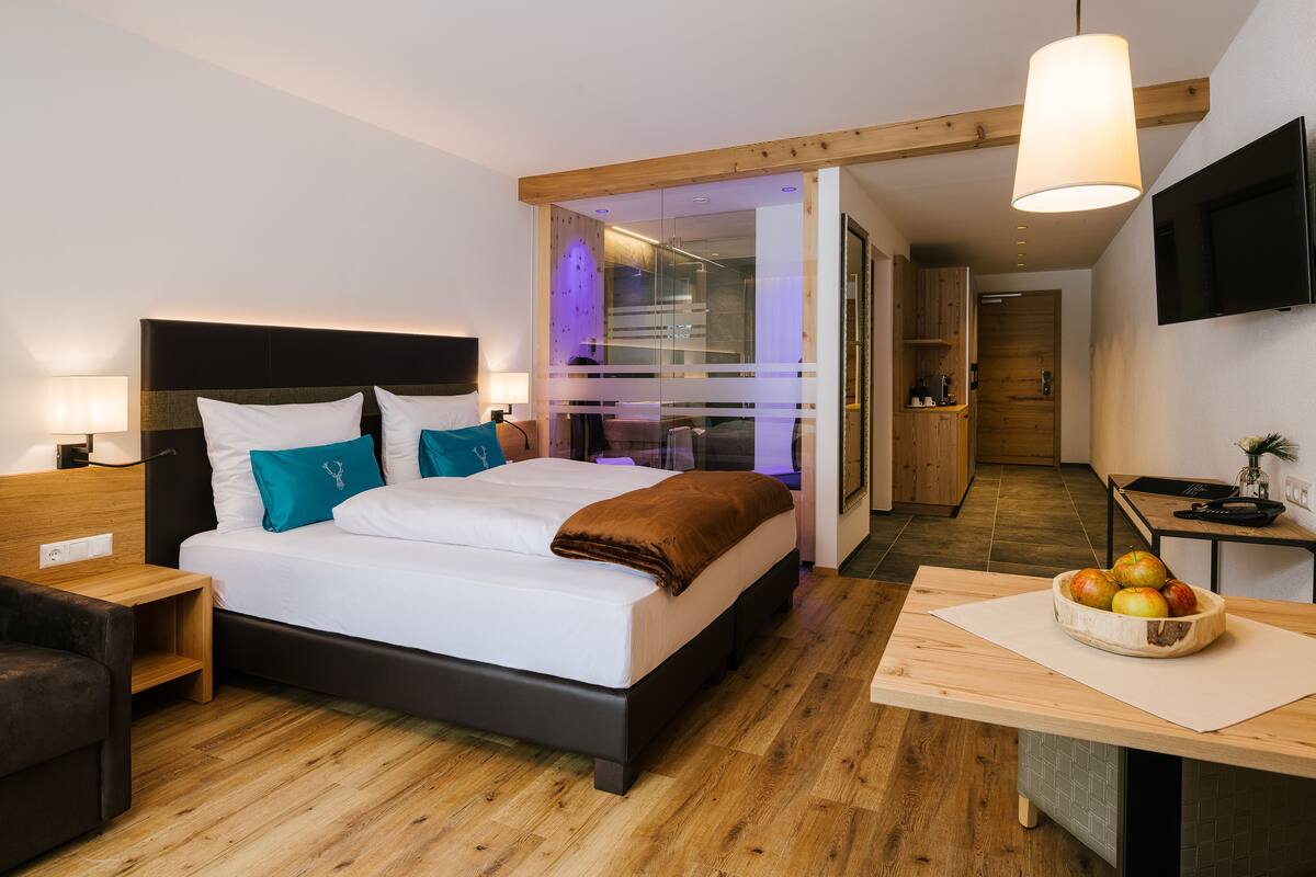 A modern hotel room with a large bed, wooden flooring, a desk with a fruit bowl, and a TV on the wall.