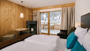 A hotel room with a wooden interior, a large bed with white and teal pillows, a small table with a fruit bowl, a fireplace, and a balcony with a view of snow-covered trees.