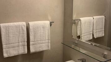 Un baño con dos toallas blancas colgadas en un perchero, un estante de vidrio sobre un lavamanos blanco y un espejo que refleja las toallas.