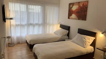 Ein Hotelzimmer mit zwei Betten, einem großen Fenster mit leichtem Vorhang, einem Bild an der Wand und einem Nachttisch mit einer Lampe.