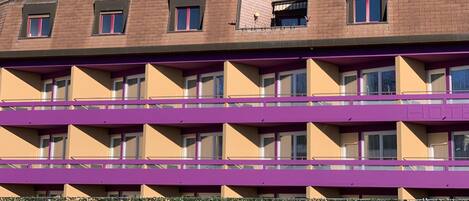 Un hotel de varios pisos con fachada morada y color crema, múltiples balcones y un estacionamiento con un automóvil.