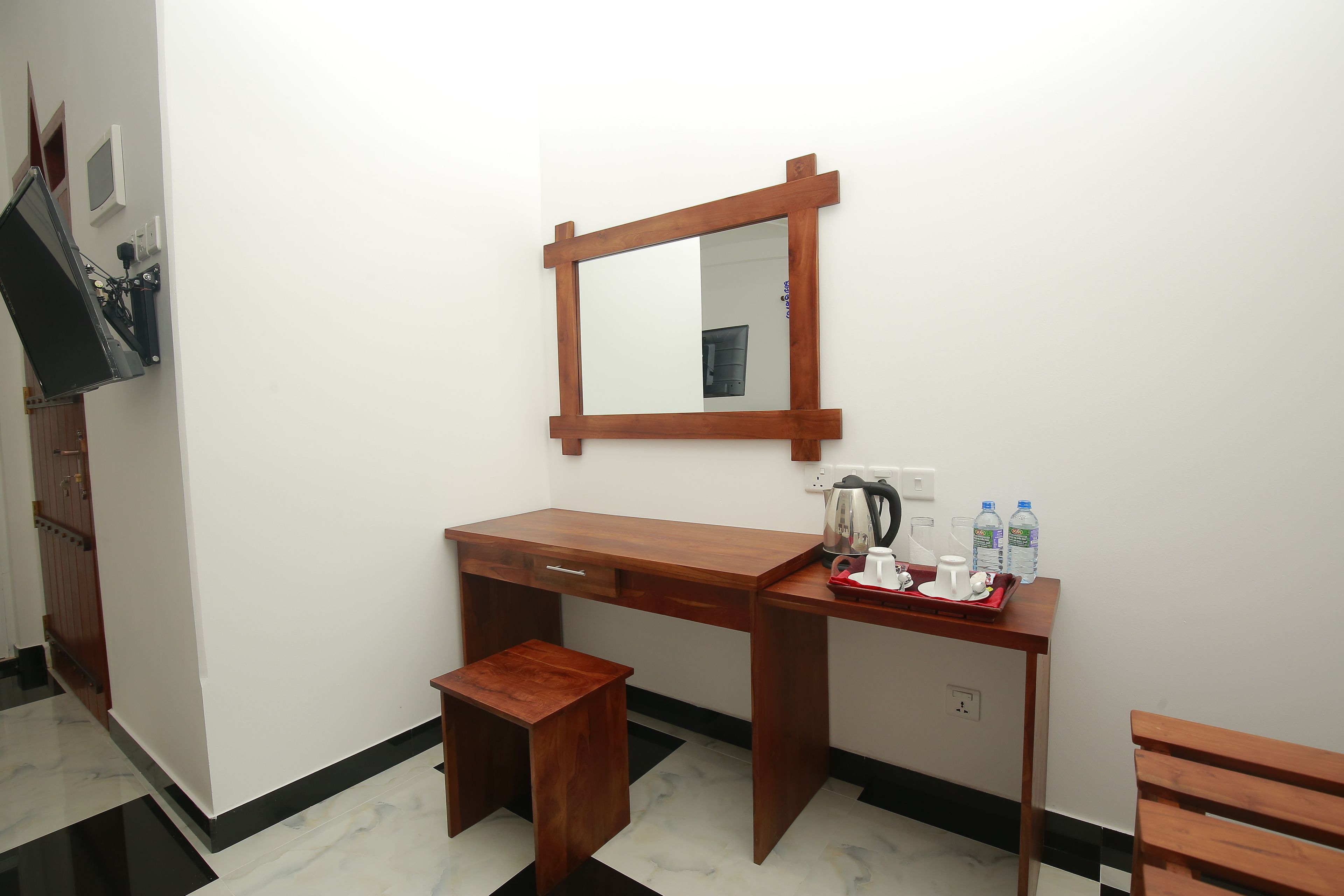 A small, wooden desk with a mirror, a small stool, and a table with a kettle, cups, and water bottles.