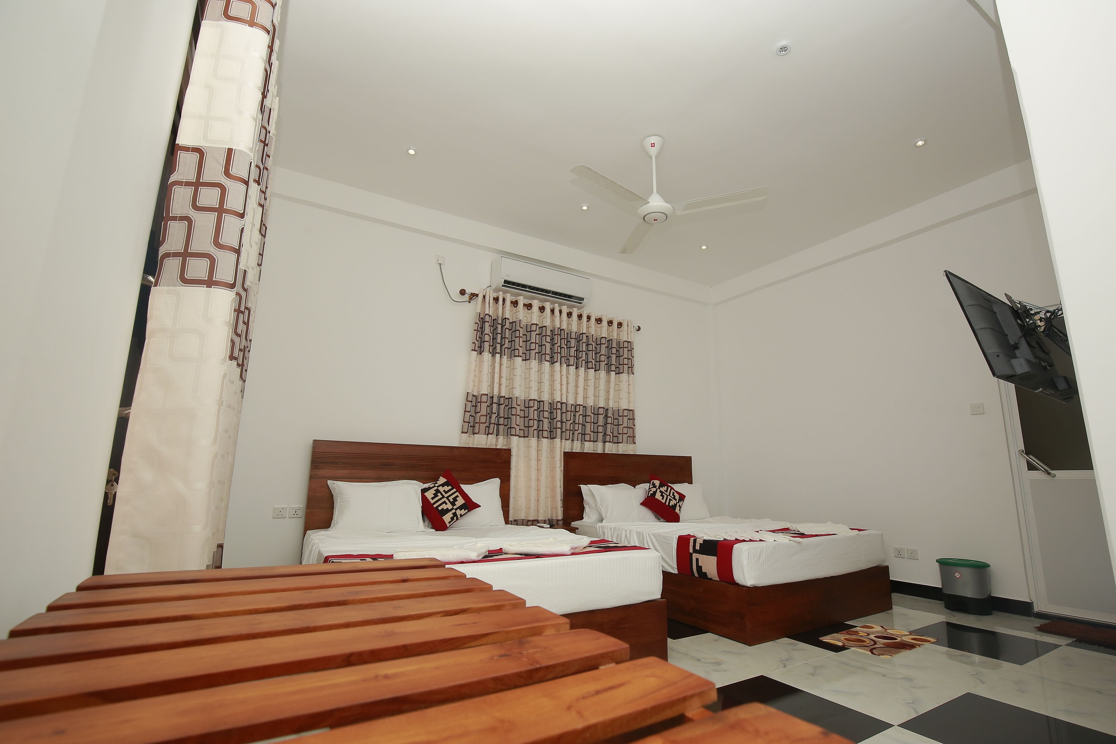 A hotel room with two beds, a ceiling fan, an air conditioning unit, a TV mounted on the wall, and a wooden bench.
