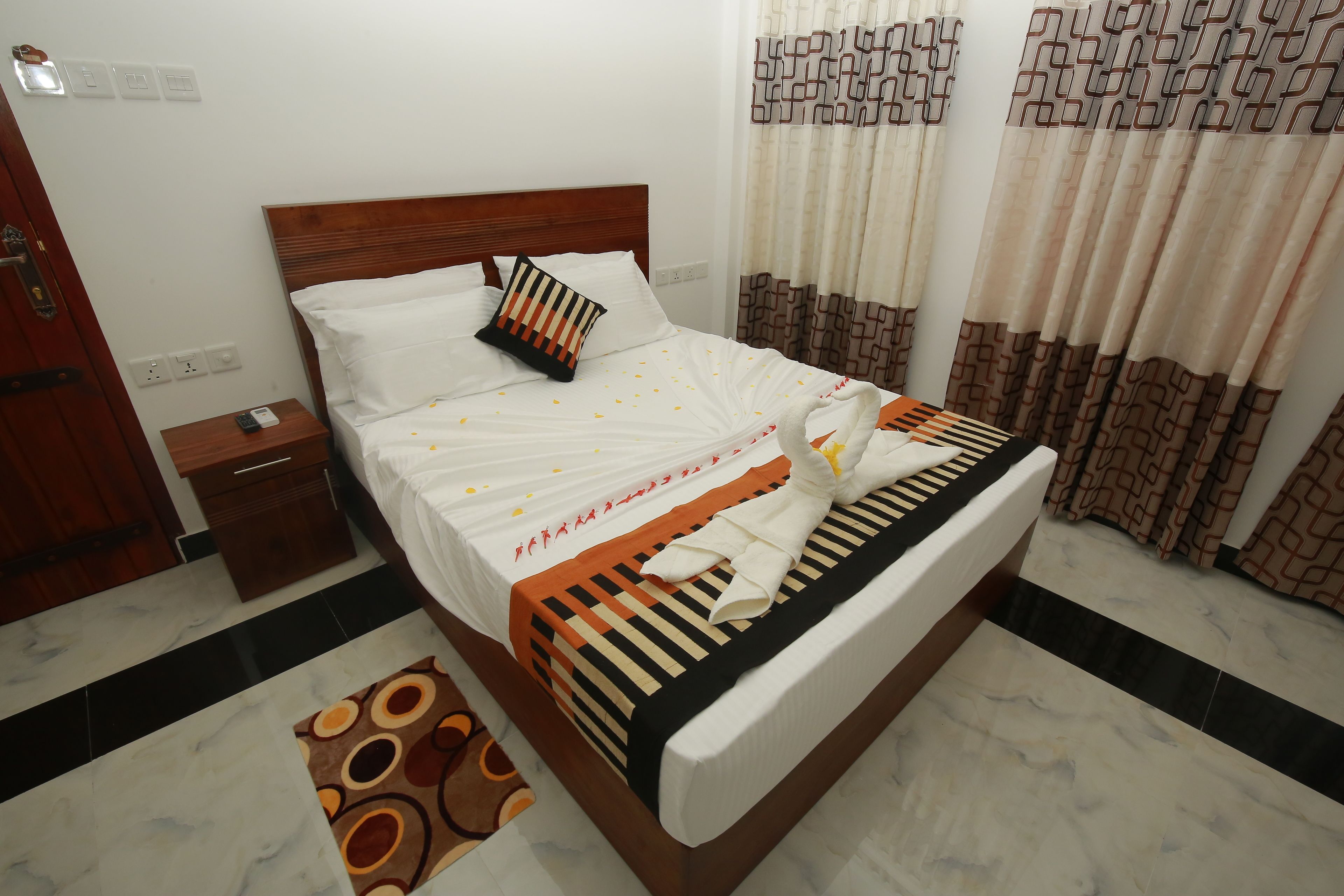 A neatly made bed with a striped bedspread, a wooden headboard, and a patterned pillow. There is a wooden nightstand with a phone and a small drawer. The room has a patterned rug and curtains with a geometric design.