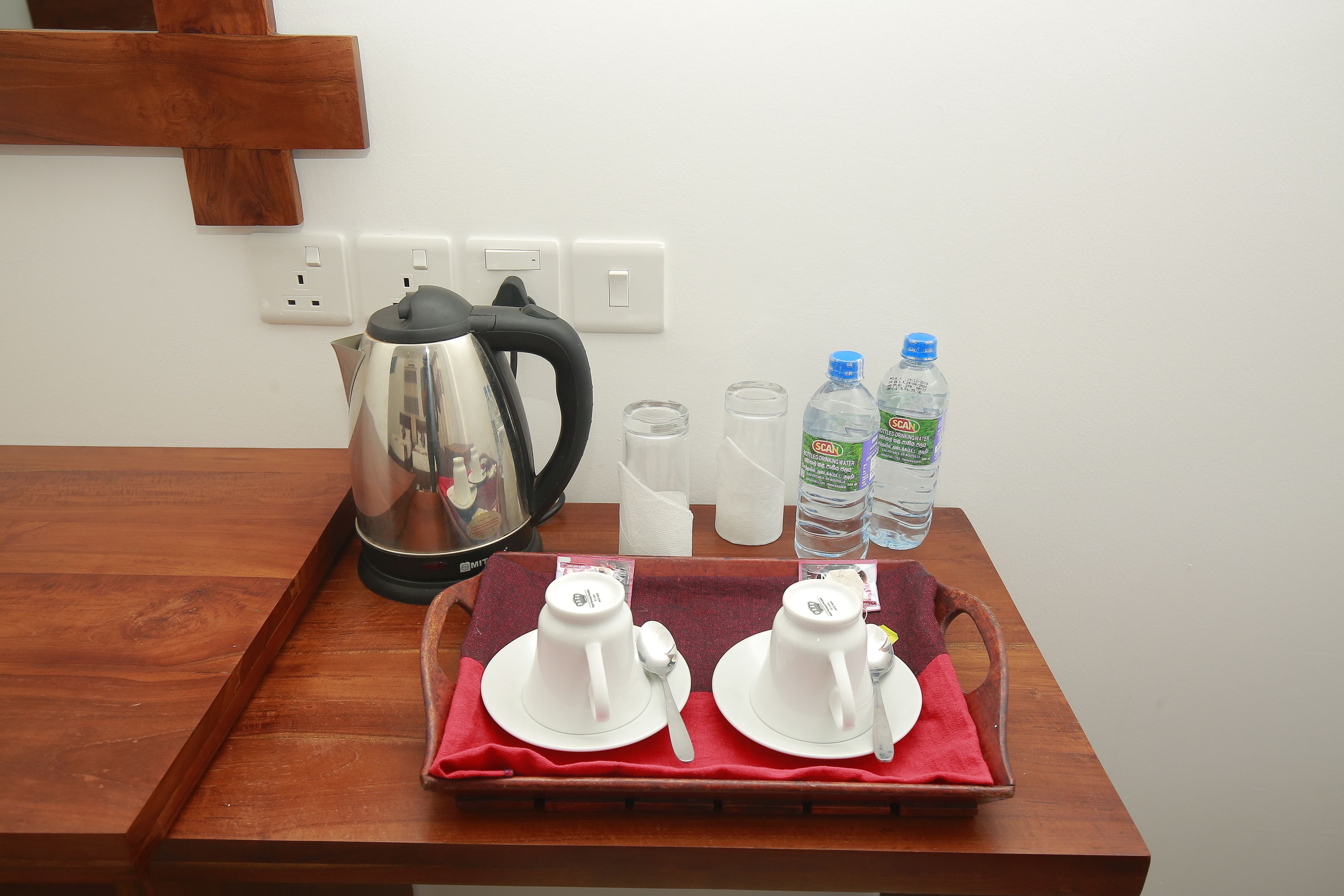 Une table en bois avec une bouilloire, deux tasses avec leurs soucoupes, un tissu rouge et deux bouteilles d’eau.