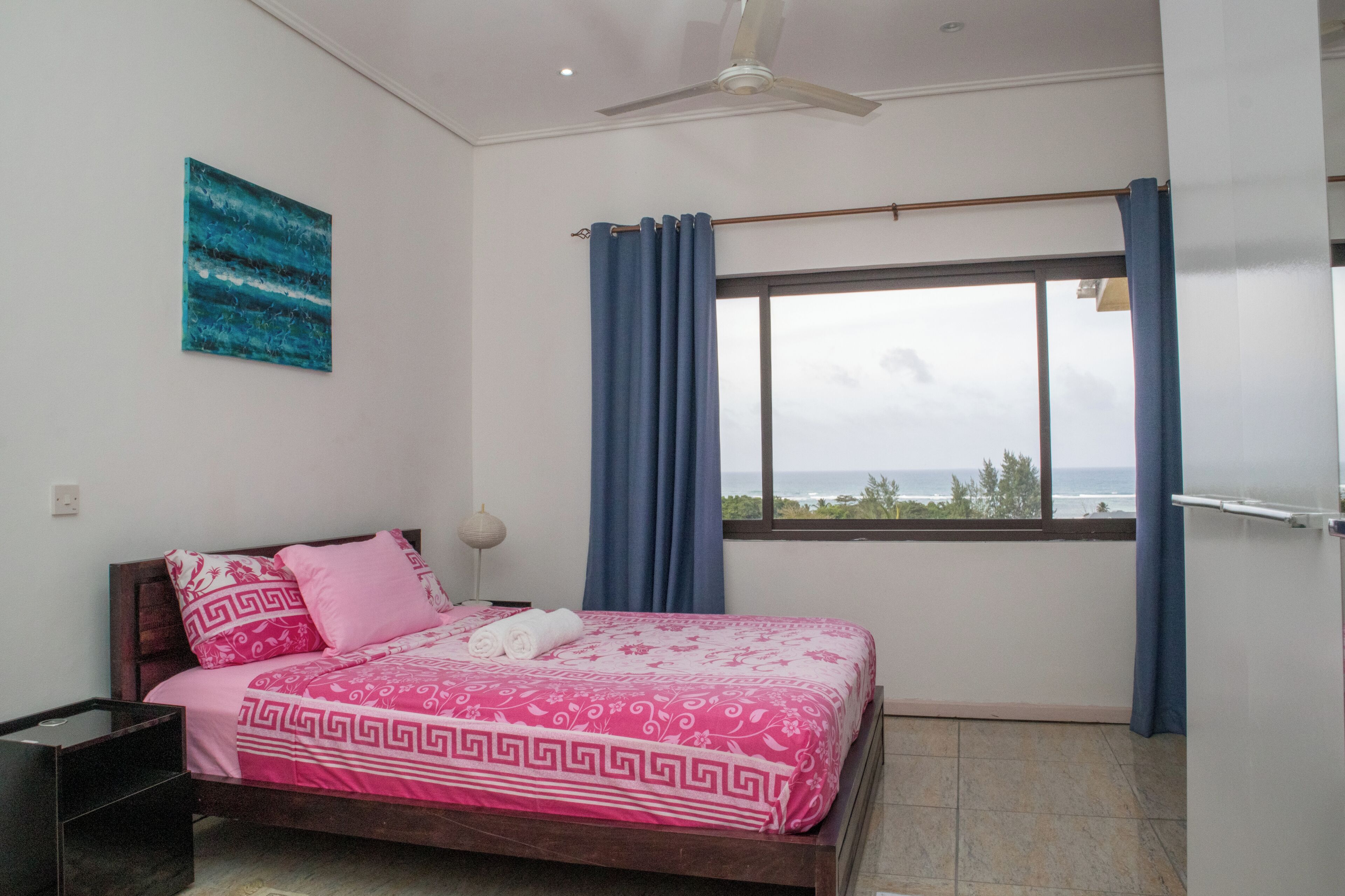 A bedroom with a bed, a ceiling fan, a window with curtains, and a view of the ocean.