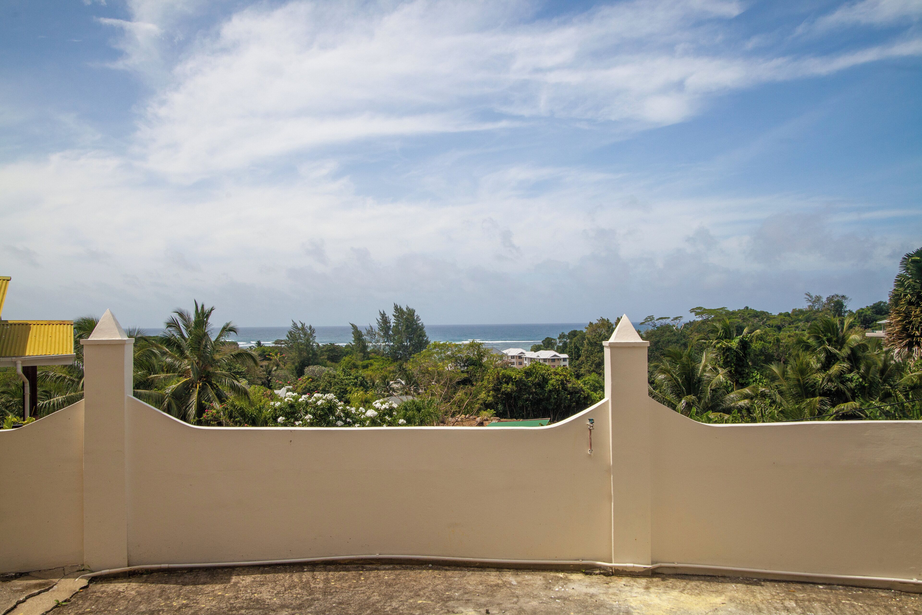 Vue depuis un balcon, avec un mur incurvé de couleur beige, donnant sur une zone côtière bordée de palmiers et où l’on aperçoit un bâtiment au loin.