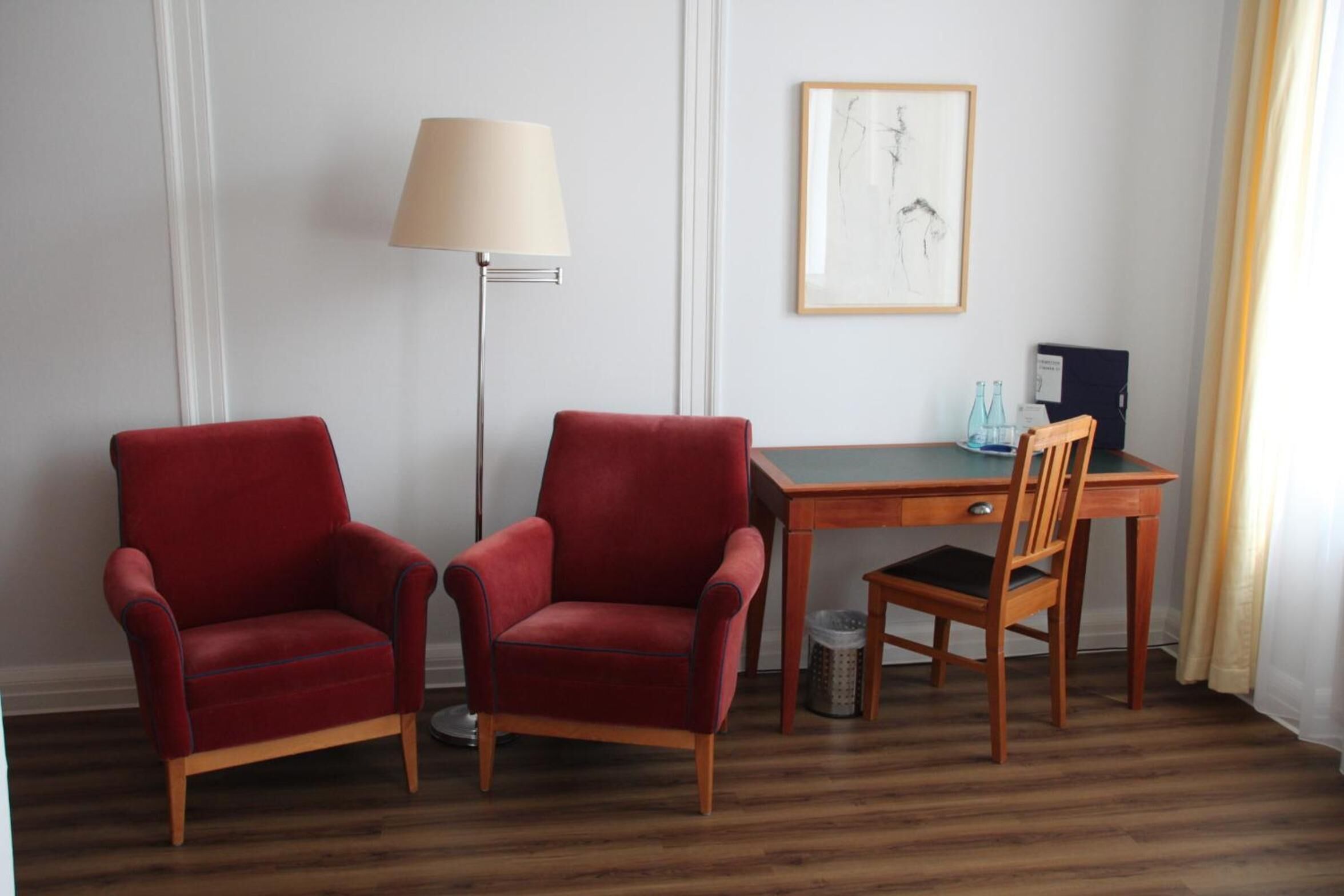 A room with two red armchairs, a wooden desk, a lamp, and a framed artwork on the wall.