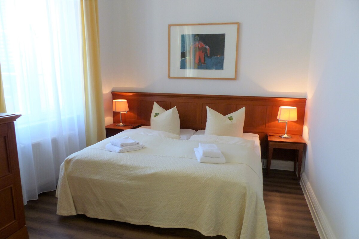 A hotel room with a large bed, two bedside tables with lamps, a framed picture on the wall, and a wooden floor.