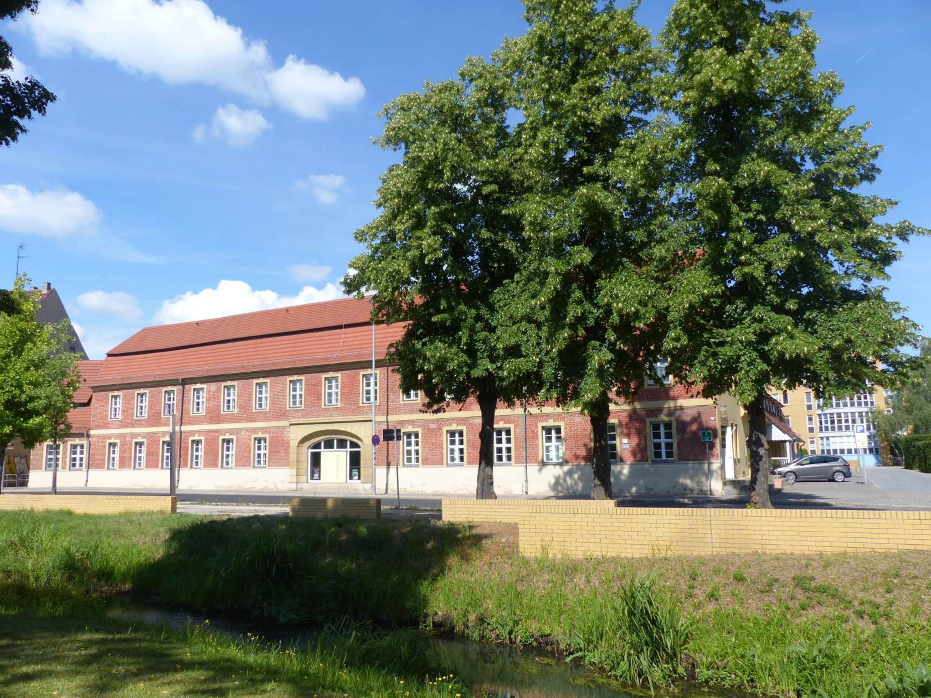 A red-brick building with a central entrance, large windows, and a surrounding grassy area with a tree.