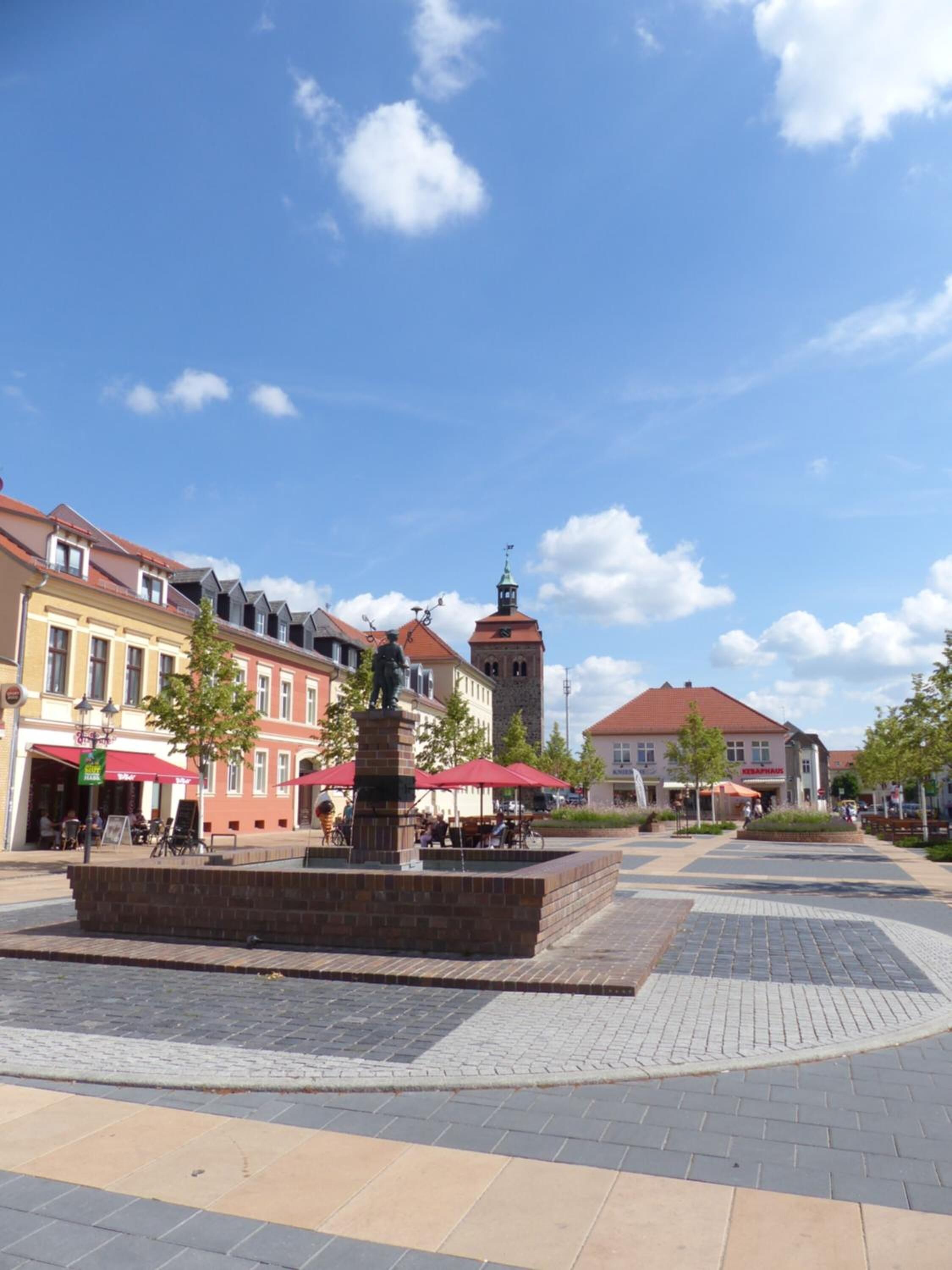 A public square with a central statue, surrounding buildings, and a clear sky.