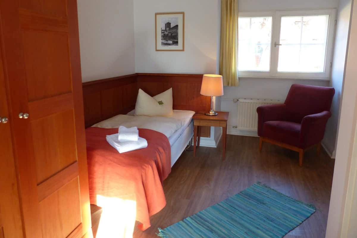 Habitación de hotel con cama, una silla, mesita de noche, lámpara, ventana con cortinas e imagen en el marco en la pared.