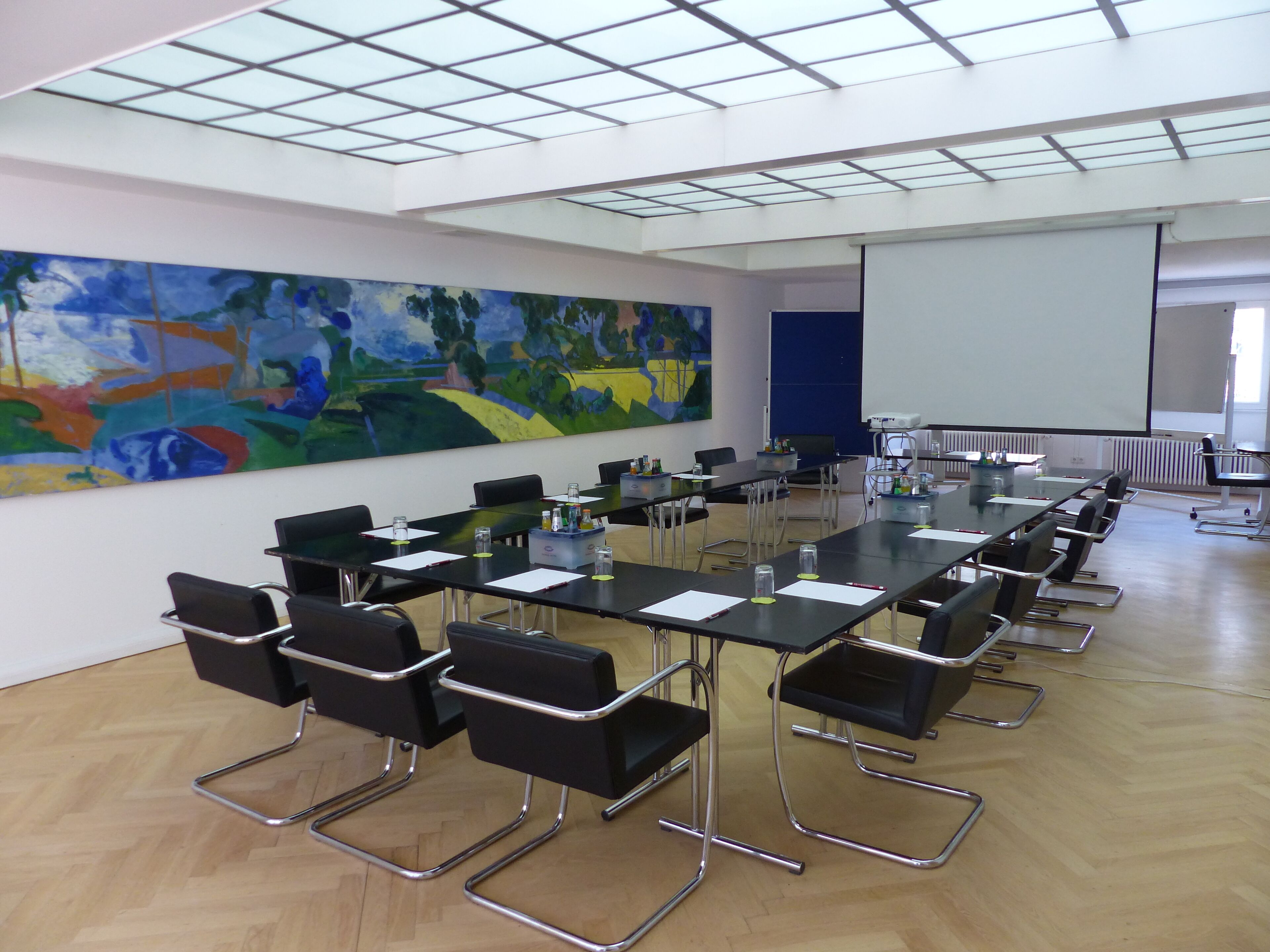 Een vergaderzaal met een lange tafel, zwarte stoelen, een projectiescherm en een groot abstract schilderij aan de muur.