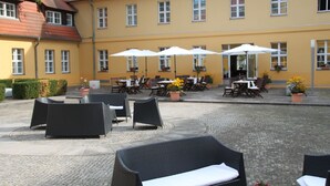 An outdoor seating area with wicker furniture and white umbrellas, set against a yellow building with multiple windows. - Hotel Vierseithof Luckenwalde (Luckenwalde)