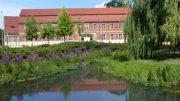 A large brick building with a red roof, surrounded by lush greenery and a pond. - Hotel Vierseithof Luckenwalde (Luckenwalde)