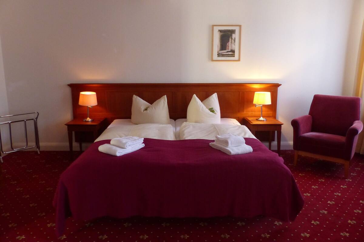 Habitación de hotel con una cama grande, dos mesitas de noche con lámparas, una silla y un cuadro en la pared.