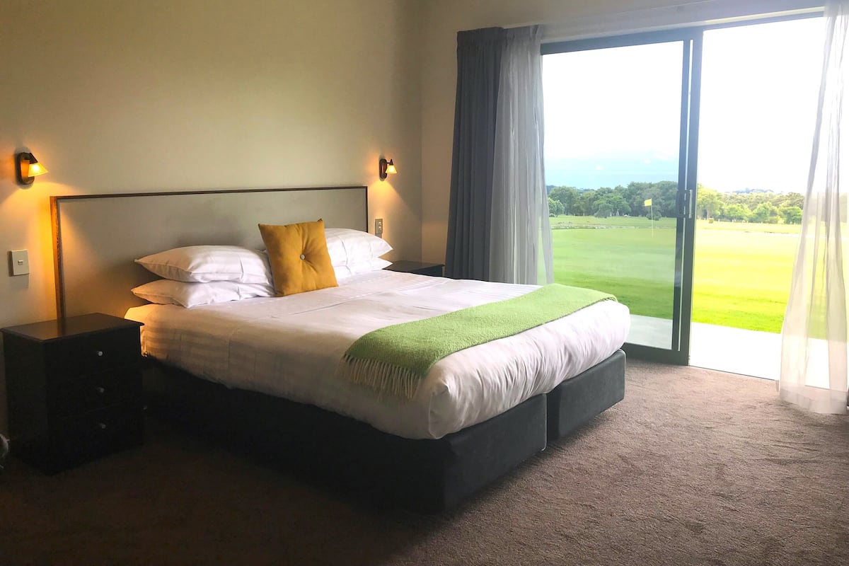 A hotel room with a large bed, two bedside tables, a view of a golf course through the window, and a green throw blanket.