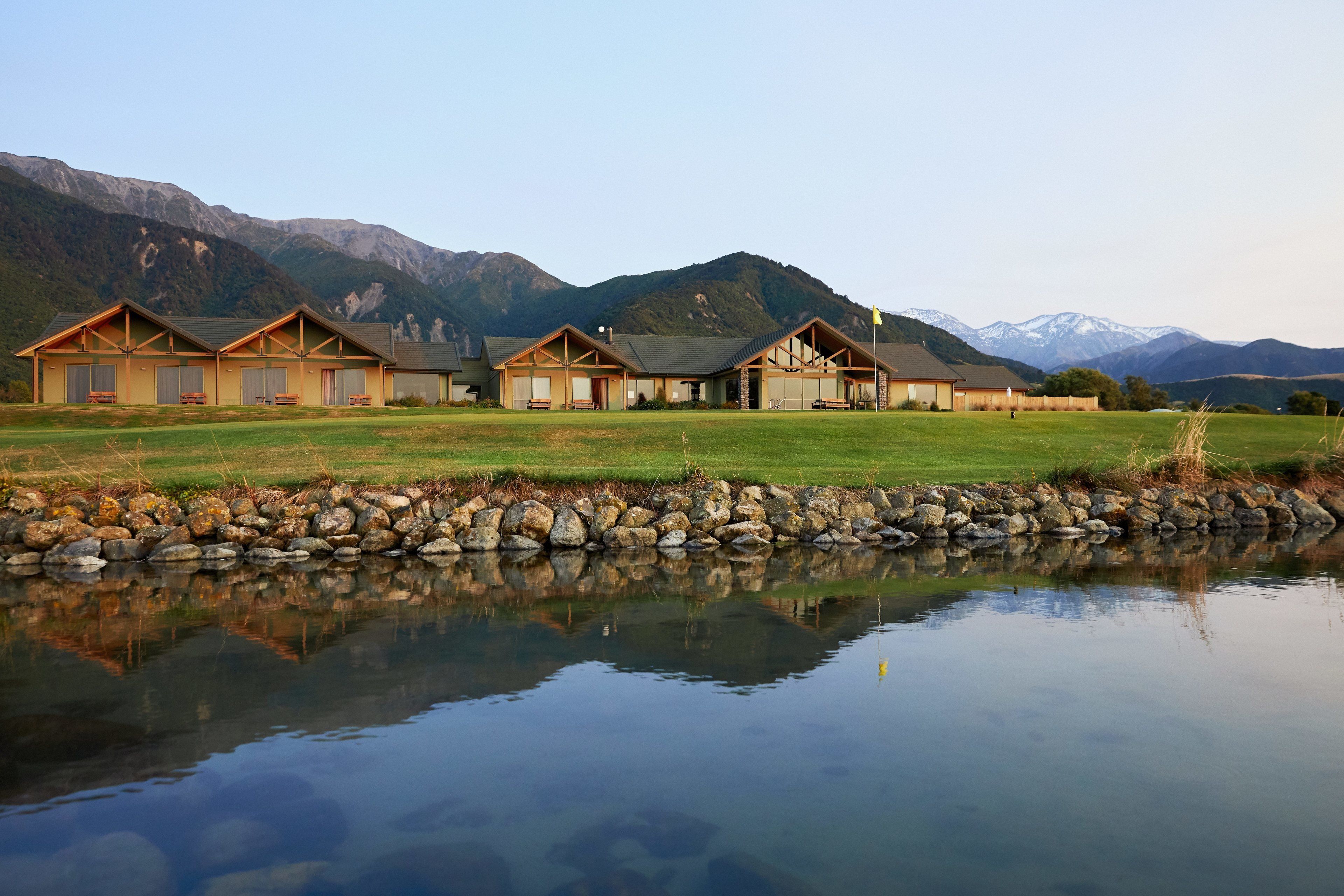 A row of log cabin-style lodges with a stone wall and a pond in front, set against a mountainous backdrop.