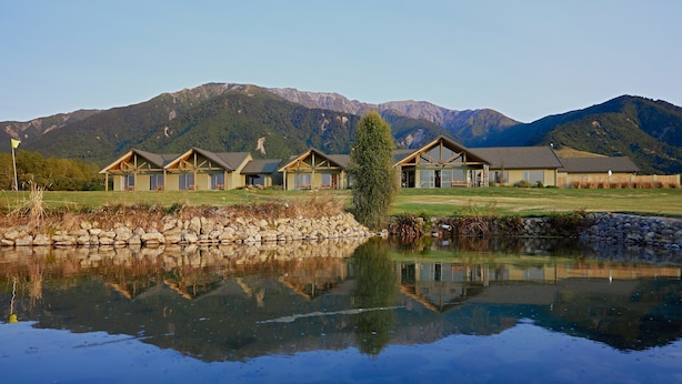 A row of log cabin-style buildings with a reflective pond in front, set against a backdrop of mountains. - Koura Bay Resort (Hapuku)