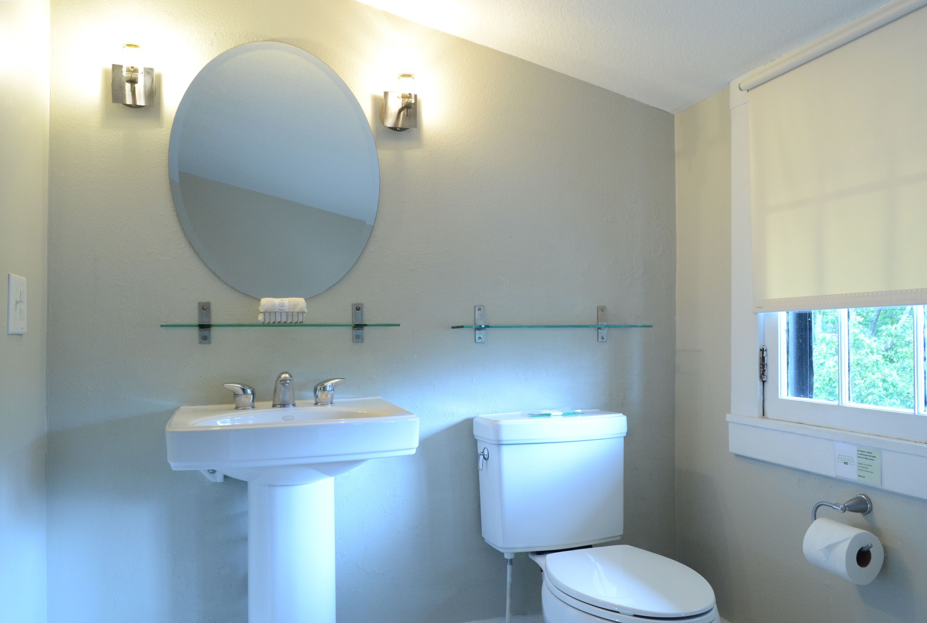 A bathroom with a white sink, a toilet, a round mirror, and a window with a view of greenery.