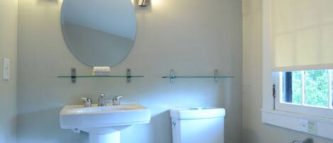 A bathroom with a white sink, a toilet, a round mirror, and a window with a view of greenery.
