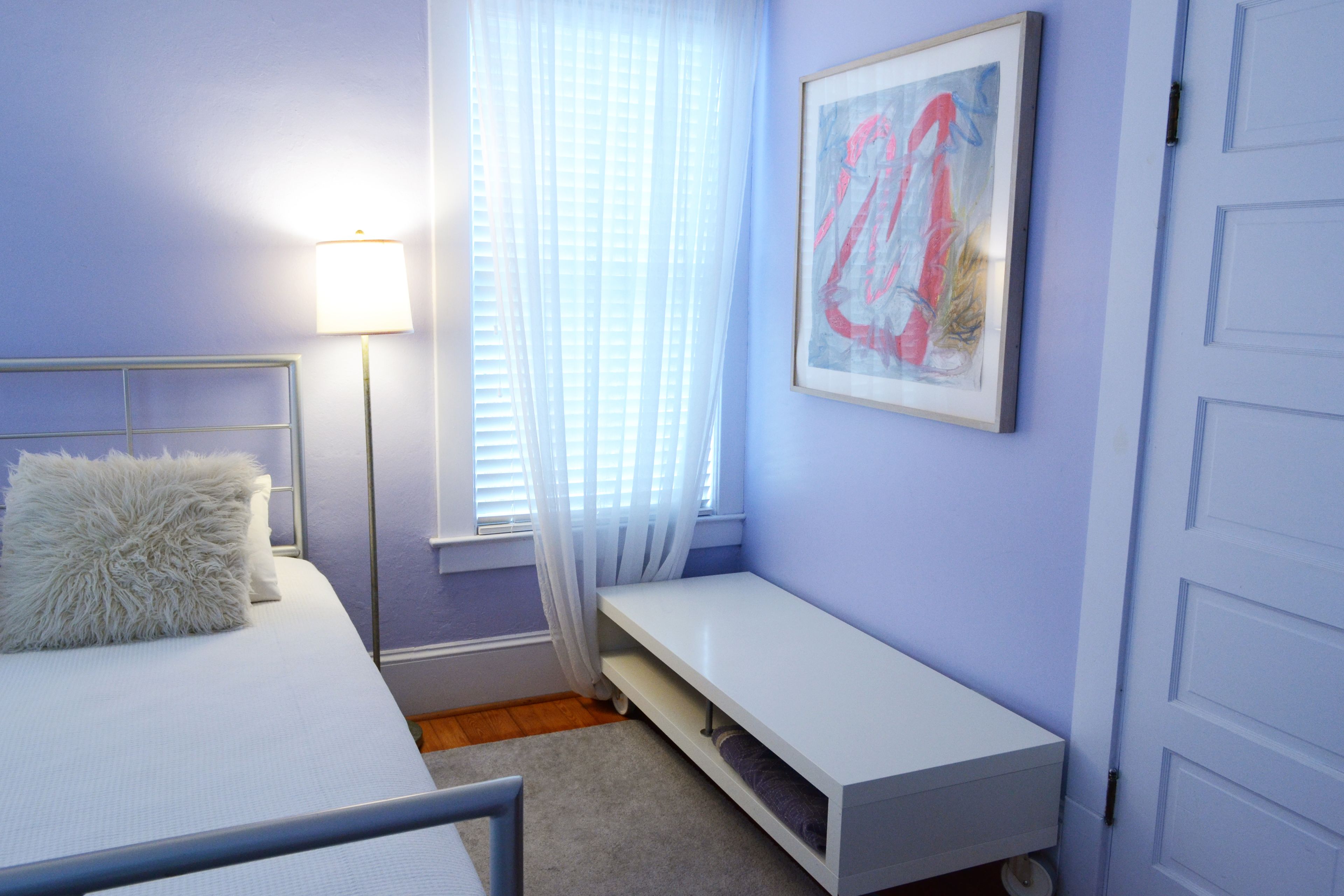 Een slaapkamer met een bed, een bankje, een lamp en een raam met gordijnen.