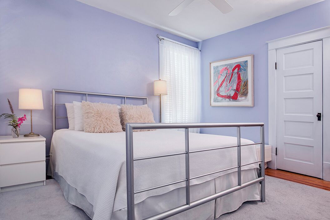 A bedroom with a metal bed frame, white bedding, a nightstand with a lamp, a framed artwork on the wall, and a white door.