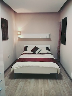 Un dormitorio con cama, mesita de noche, estante en la pared y luz de pared.