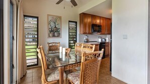 A dining area with a glass table and wicker chairs, a ceiling fan, wooden cabinets, and a kitchen in the background.