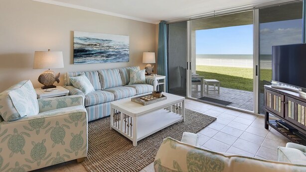 A living room with a sofa, coffee table, and a television. There is a large window with a view of the ocean.