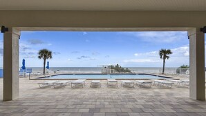 An outdoor swimming pool area with lounge chairs and palm trees, overlooking the sea.