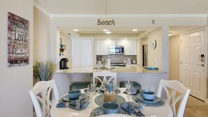 A dining area with a white table set for four, surrounded by white chairs. A kitchen with white cabinets and a microwave is visible in the background.