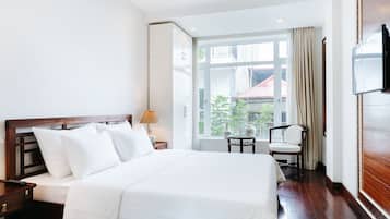 Habitación de hotel con una cama grande, una mesita de noche, una lámpara, una silla y una ventana con vistas a edificios y árboles.