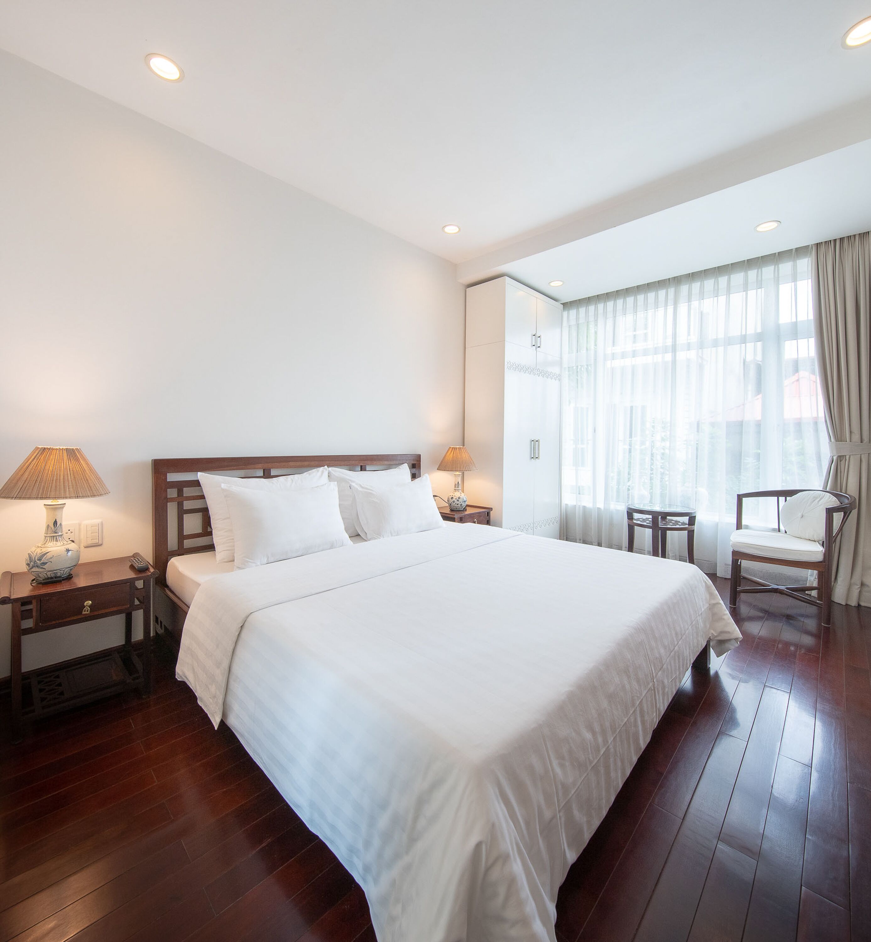Habitación de hotel con una cama grande, dos mesitas de noche con lámparas, una silla y un amplio ventanal con cortinas.