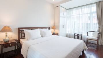 Habitación de hotel con una cama grande, dos mesitas de noche con lámparas, una silla y un amplio ventanal con cortinas.