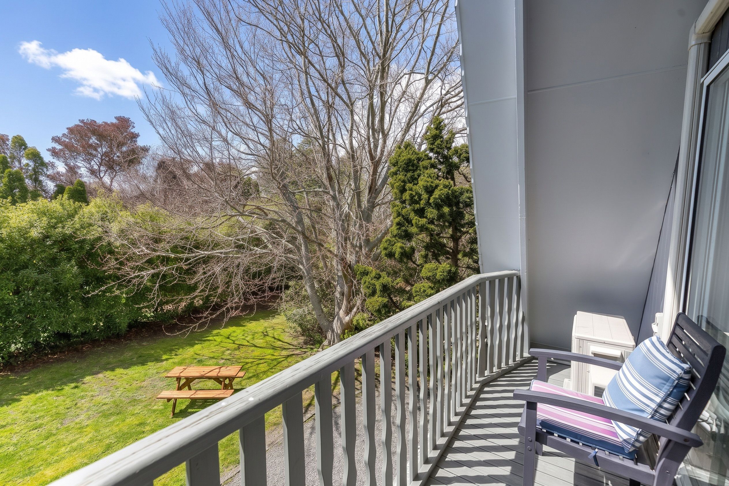 Un balcon avec un banc en bois et une table de pique-nique, donnant sur un jardin où l’on peut admirer différentes espèces d’arbres et d’arbustes.