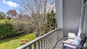 A balcony with a wooden bench and a picnic table, overlooking a garden with various trees and shrubs.