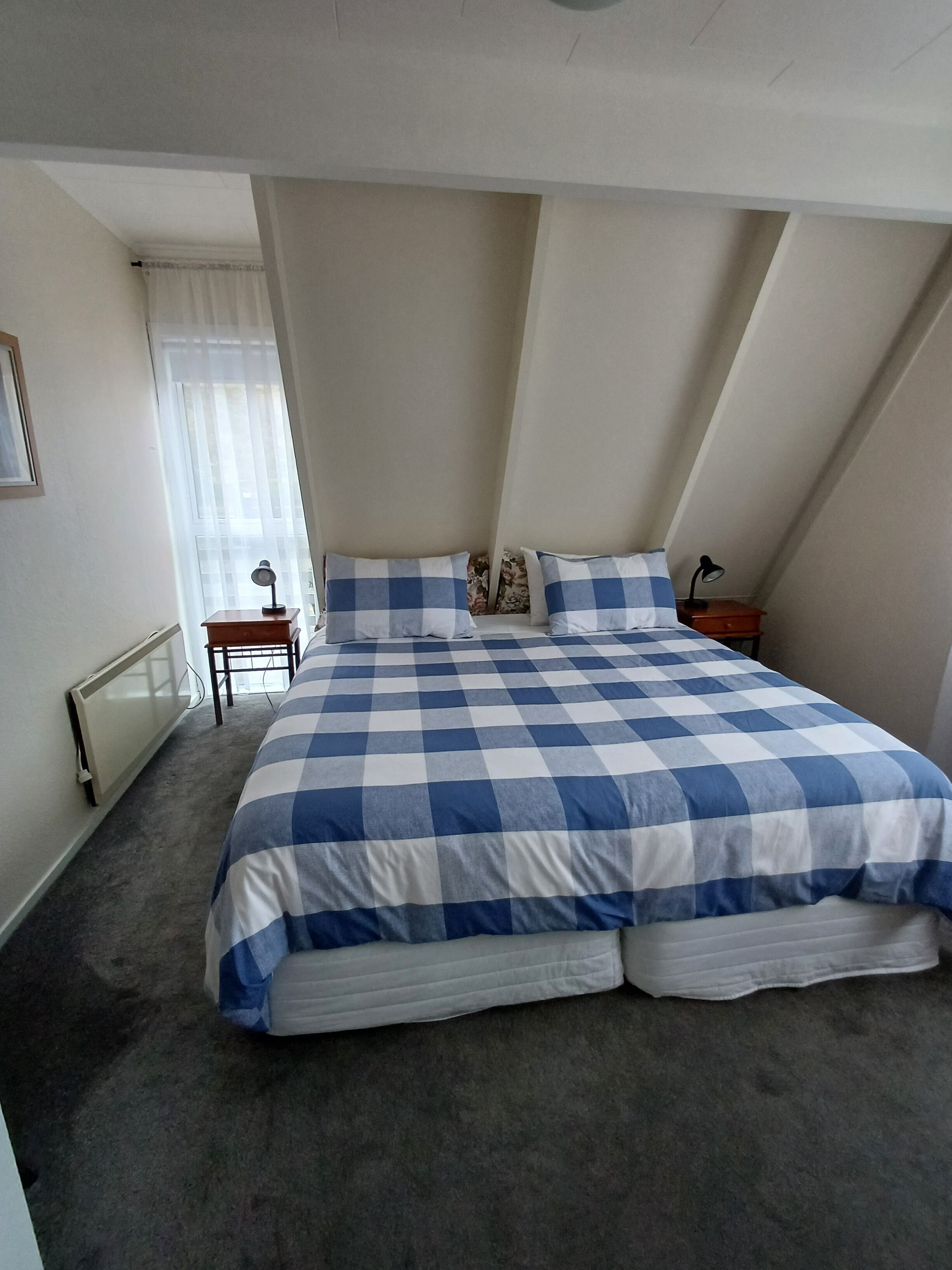 A bedroom with a large bed, a nightstand, a lamp, and a window with sheer curtains.